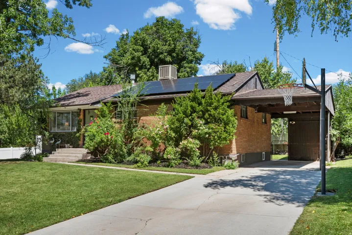 Brick exterior with solar panels, covered carport, concrete driveway, and mature landscaping