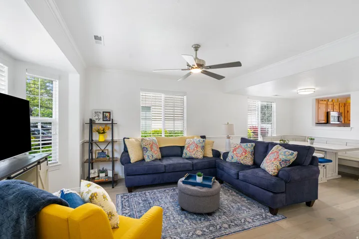 Bright open-concept living area featuring wood-finish flooring, multiple windows with blinds, a ceiling fan, and crown molding
