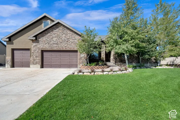 Stucco and stone exterior with a two-car garage, concrete driveway, front lawn, and mature trees