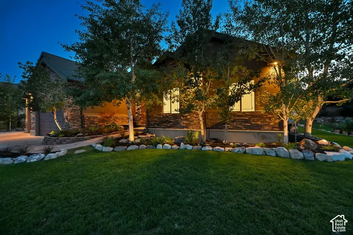 Stone facade with integrated outdoor lighting, landscaped grounds with mature trees, and a paved driveway