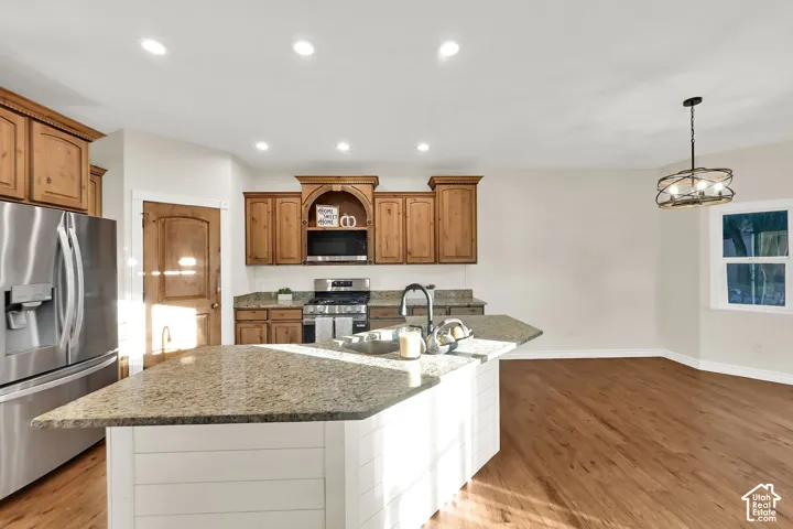 Kitchen featuring wood cabinetry, stainless steel appliances, a granite-finish island with a sink, wood-finish flooring, and recessed lighting