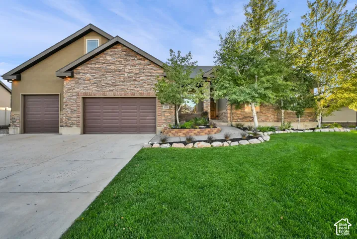 Stone facade with stucco accents, two-car garage, concrete driveway, and a landscaped front yard with mature trees
