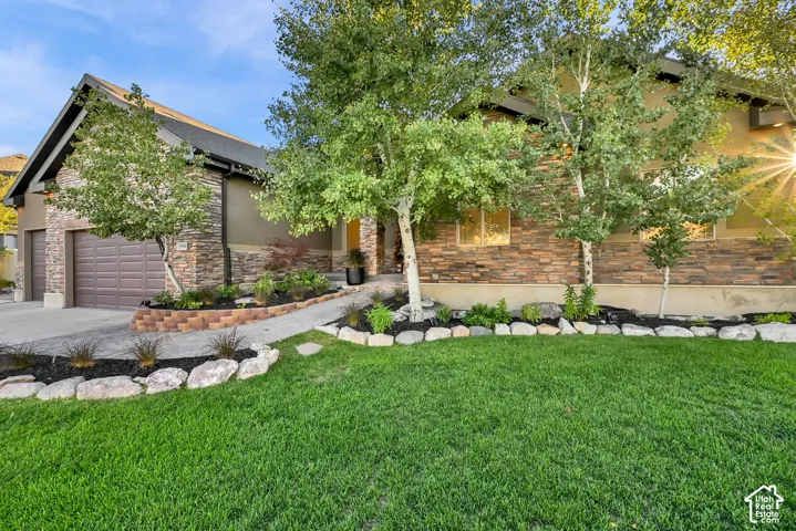 Stone and stucco exterior with multiple mature trees, a manicured lawn, and an integrated two-car garage