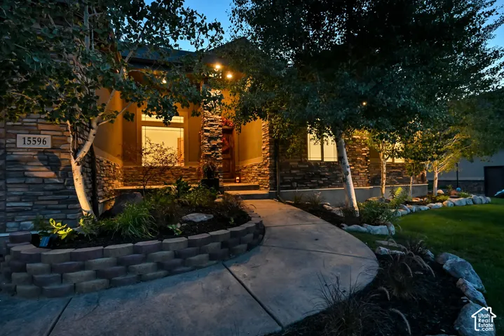 Stone facade with stucco accents, curved concrete walkway, landscaped garden beds with block retaining wall, mature trees, and exterior lighting