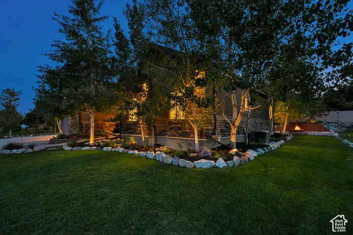 Stucco and stone exterior, manicured lawn, established trees, landscape lighting, and rock-edged garden beds