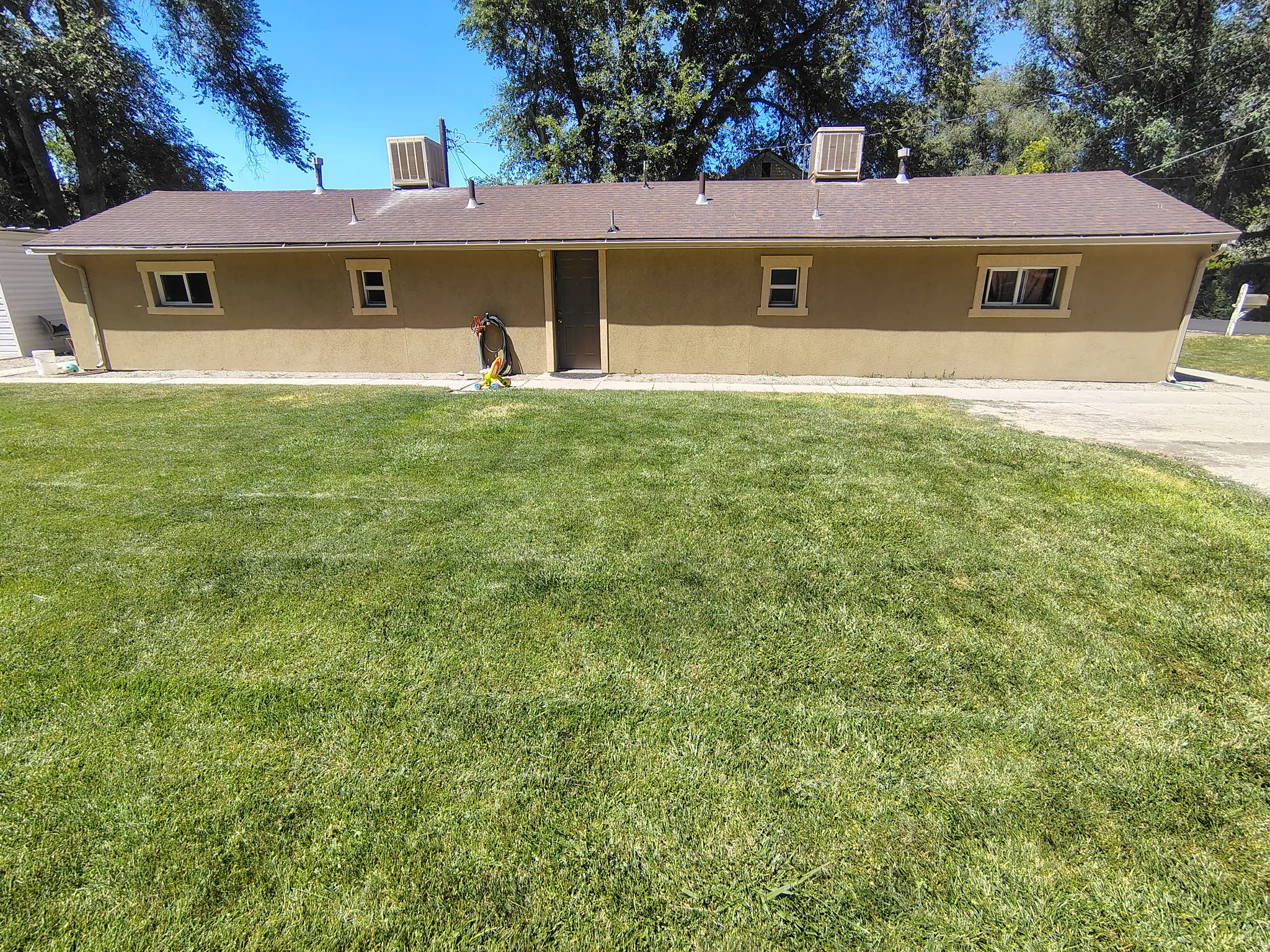 Expansive green lawn with a stucco exterior structure featuring multiple windows, a central entry door, and a brown shingle roof