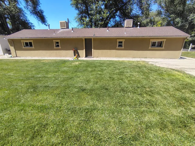 Expansive green lawn with a stucco exterior structure featuring multiple windows, a central entry door, and a brown shingle roof