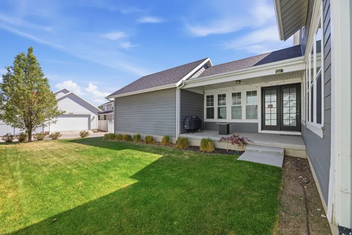 Expansive green lawn, covered concrete patio, black French doors with grilles, multiple windows with white trim, and gray horizontal siding