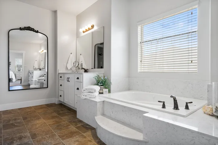 Soaking tub with built-in steps, expansive vanity with white cabinetry, large window with horizontal blinds, and tile flooring