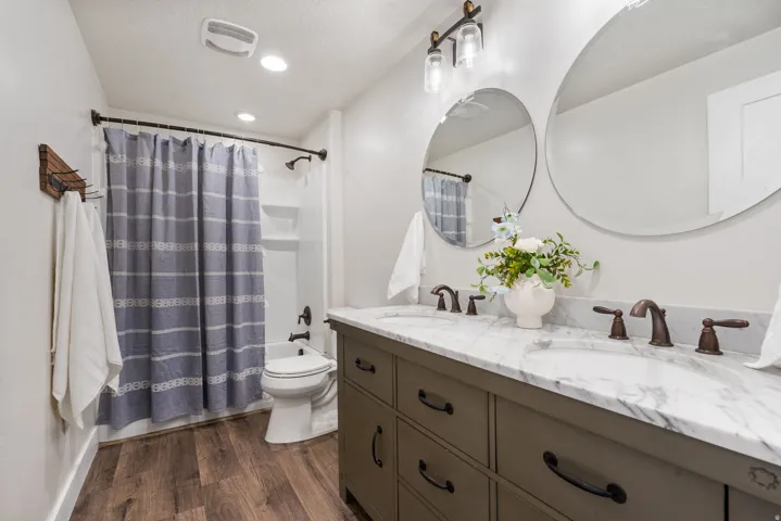 Dual round vanity mirrors, double sink vanity with stone countertop, wood-finish flooring, and recessed lighting