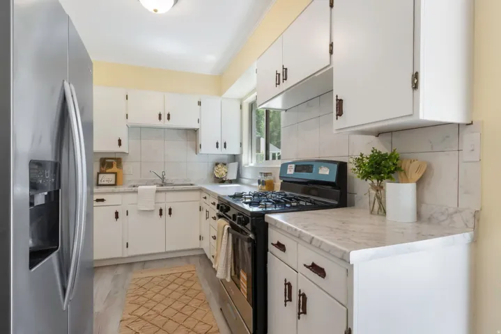 White cabinetry with dark hardware, stainless steel refrigerator, gas range, light-toned wood-finish flooring, and a window for natural light