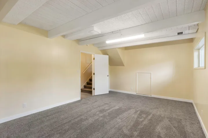 Recessed ceiling beams, textured ceiling, light-toned walls, gray-toned carpeting, and a visible staircase