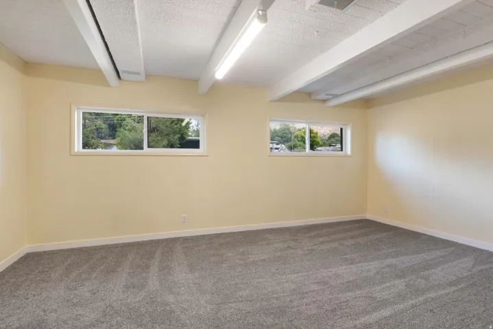 Exposed ceiling beams, recessed lighting, neutral wall paint, gray carpeting, and two horizontally oriented windows