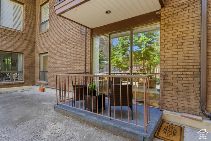 Covered patio with brick exterior walls, large multi-panel glass doors and windows framing mature landscaping, concrete flooring, and metal safety railing