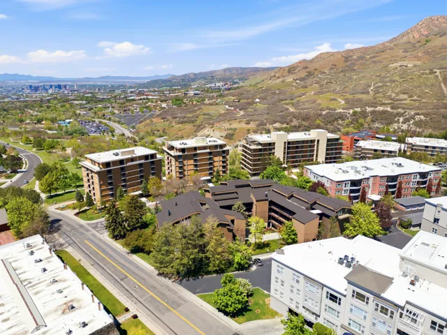 Apartment buildings with multiple balconies, surrounded by mature trees and manicured landscaping, against a backdrop of distant mountains and a city skyline