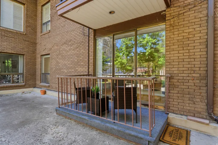 Covered patio with brick exterior walls, large multi-panel glass doors and windows framing mature landscaping, concrete flooring, and metal safety railing