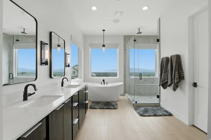Double vanity with integrated sinks, freestanding soaking tub, glass-enclosed shower, light wood-finish flooring, and contemporary pendant lighting