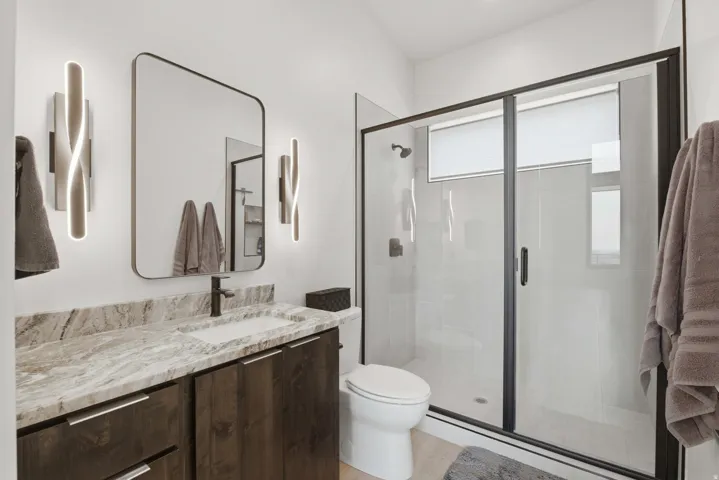 Custom vanity with stone countertop and integrated sink, modern wall sconces, large framed mirror, and a glass-enclosed shower with dark trim