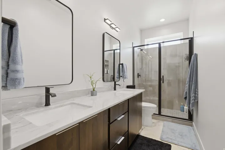 Dual undermount sinks with matte black faucets, a spacious floating vanity with wood-finish cabinetry, and a walk-in shower with clear glass enclosure and dark trim