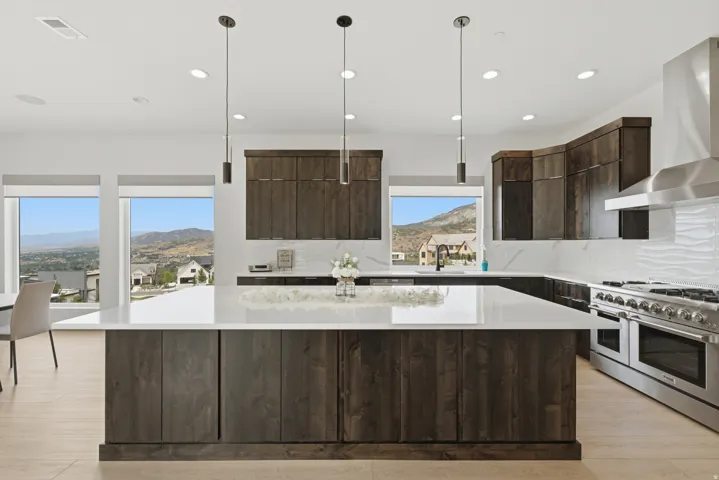 Spacious kitchen featuring an expansive central island with a white solid surface countertop, dark wood-finish cabinetry, a large stainless steel range with a professional-grade hood, and multiple windows showcasing a mountainous landscape