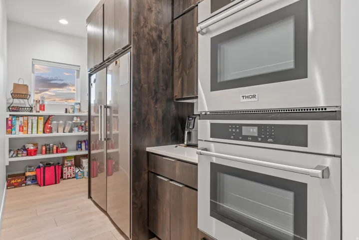 Integrated stainless steel double ovens, sleek stainless steel refrigerator, dark wood-finish cabinetry, light wood-finish flooring, and a dedicated pantry space