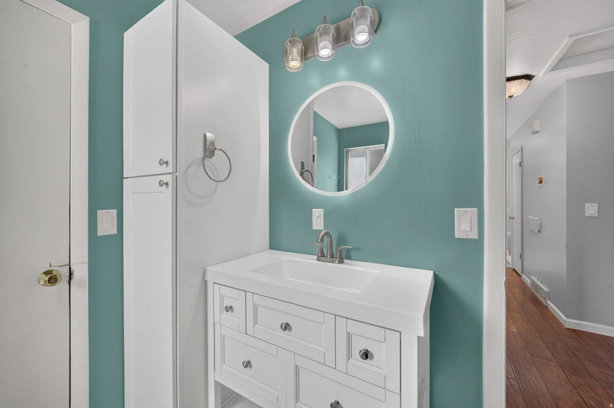 White vanity with an integrated sink and brushed nickel faucet, illuminated LED-backlit circular mirror, and overhead tri-bulb light fixture