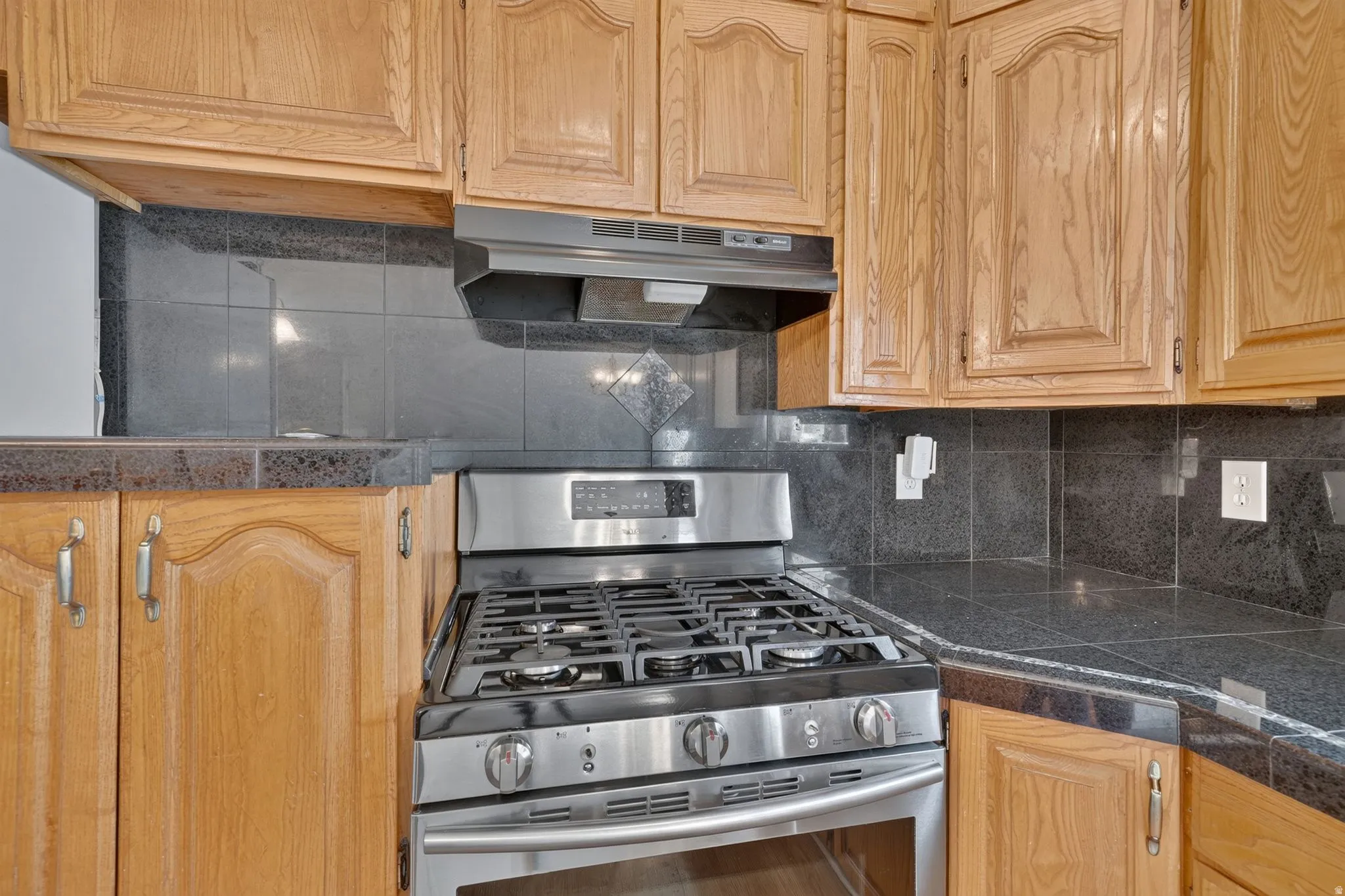 Wood-finish cabinetry, stainless steel gas range, coordinating range hood, and dark stone-finish countertops with a tiled backsplash