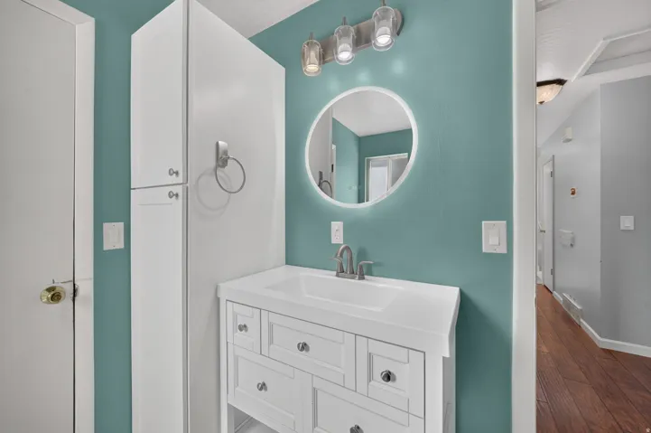 White vanity with an integrated sink and brushed nickel faucet, illuminated LED-backlit circular mirror, and overhead tri-bulb light fixture