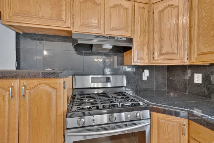 Wood-finish cabinetry, stainless steel gas range, coordinating range hood, and dark stone-finish countertops with a tiled backsplash