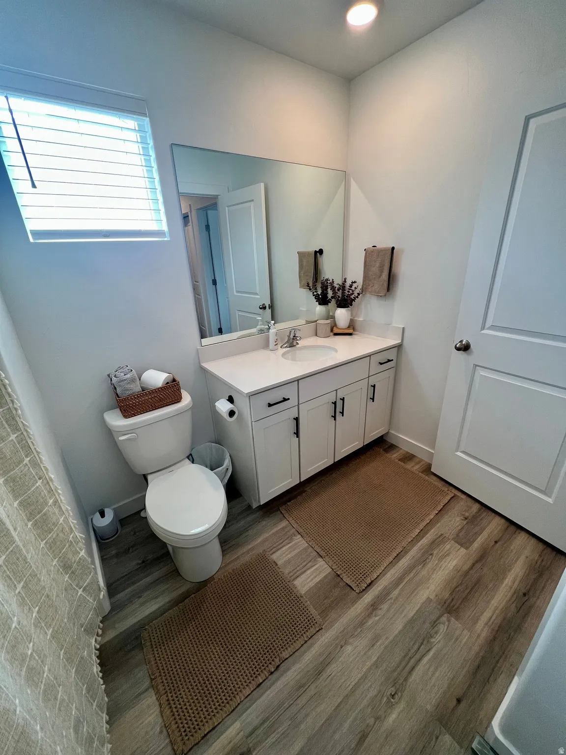 White vanity with an integrated sink, large wall-mounted mirror, wood-finish flooring, and a window with blinds