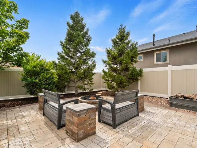 Paved patio area featuring a built-in fire pit with stone surround, complemented by mature trees and shrubs, and a privacy fence