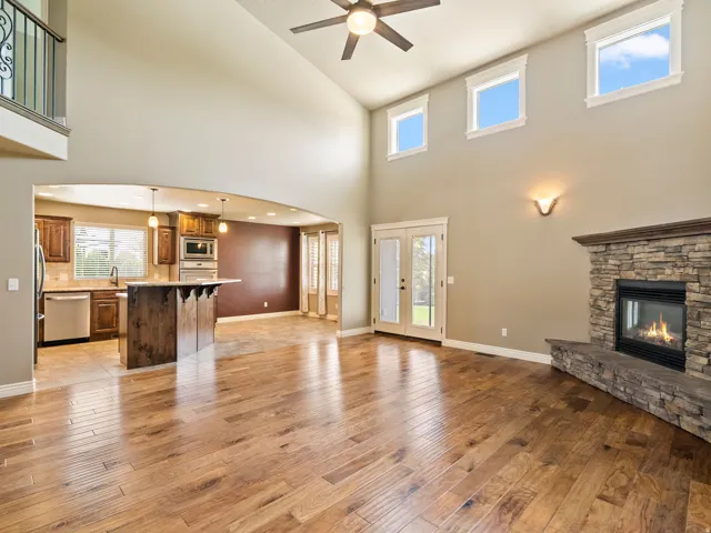 Vaulted ceiling with ceiling fan, wood-finish flooring, stone-clad fireplace, French doors, and upper transom windows