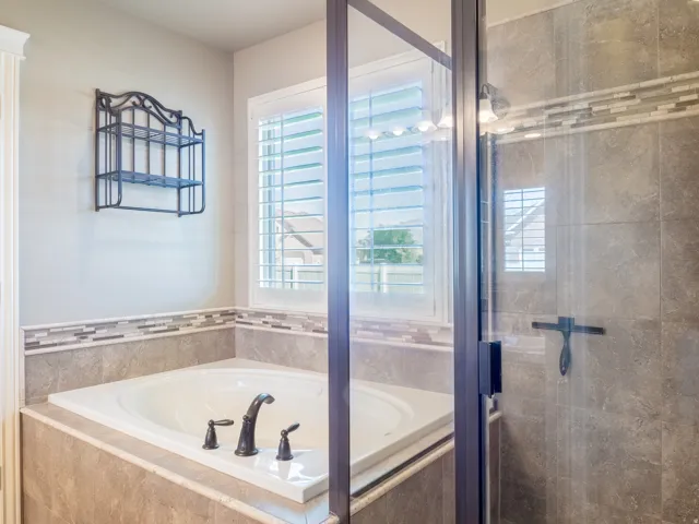 Built-in soaking tub with dark-toned fixtures, tiled surround, and plantation shutters