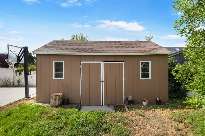 Brown storage shed with a shingle roof, double front doors, and two windows