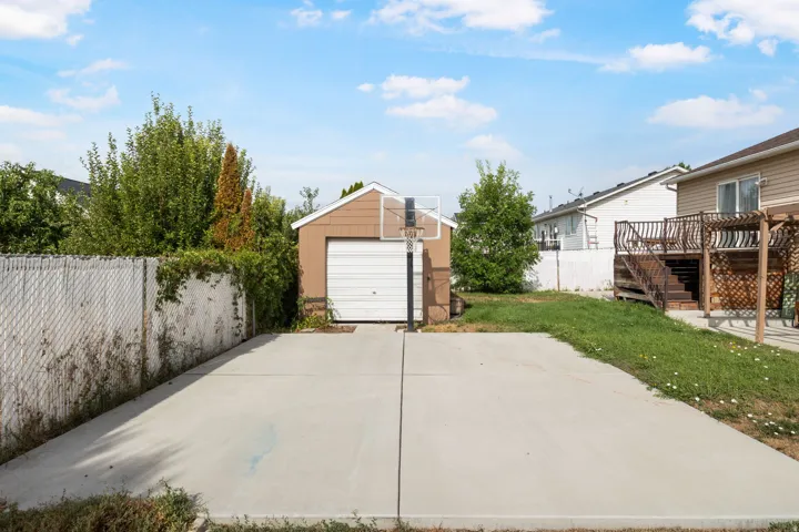 Detached shed with garage door access, paved concrete pad, chain link fencing, mature trees and shrubs, and a wood-decked patio with lattice and pergola