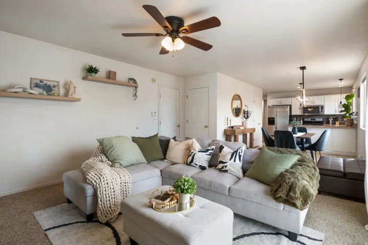 Open-concept living area with a ceiling fan, floating wall shelves, and an adjacent kitchen featuring white cabinetry and pendant lighting