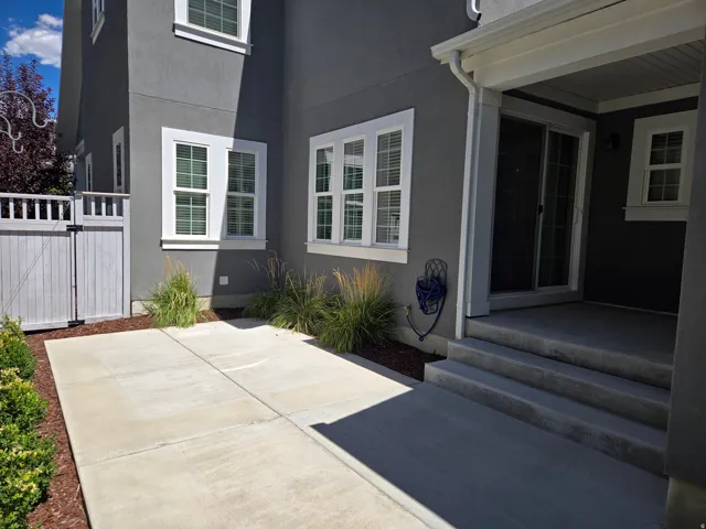 Exterior featuring a grey stucco finish, white-framed windows, a white privacy fence with gate, concrete patio, and mature landscaping