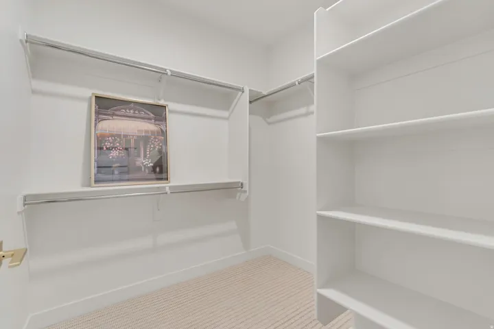 Walk-in closet featuring white built-in shelving, two levels of hanging rods, and light-toned carpet flooring