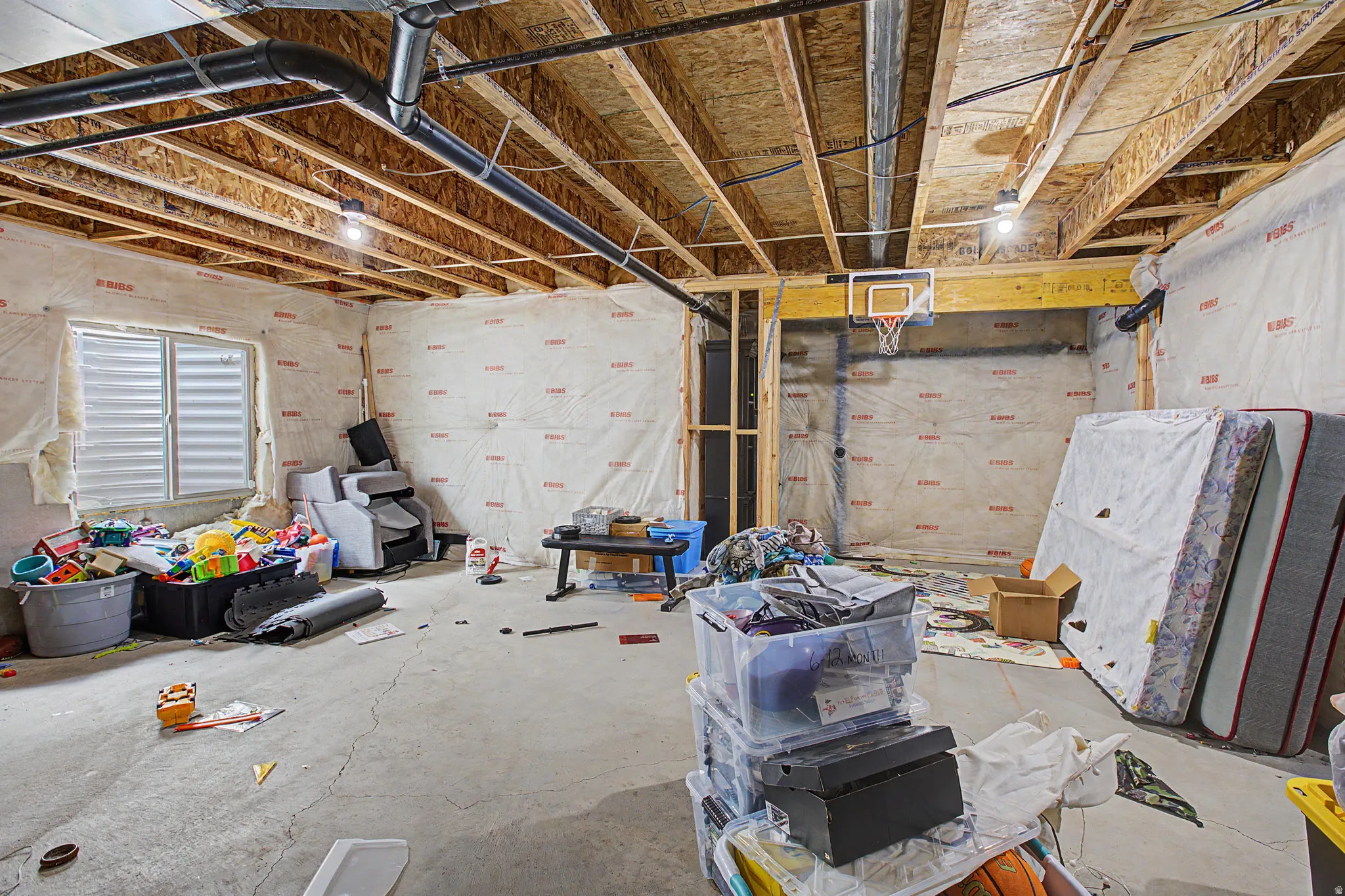 Unfinished basement space with exposed wood joists, concrete flooring, visible plumbing, and an egress window