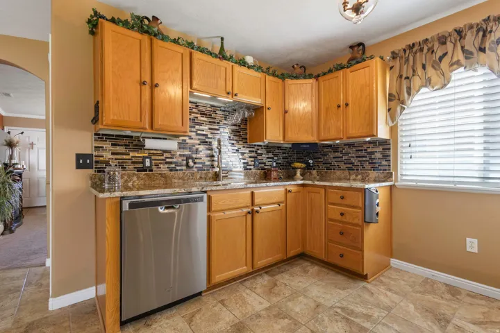 Wood-finish cabinetry, mosaic tile backsplash, stainless steel dishwasher, and a window with blinds