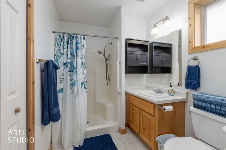 Built-in shower with grab bar, single vanity with wood-finish cabinetry, countertop with integrated sink, wall-mounted mirror, and wood-framed window