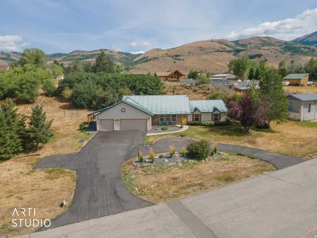 Two-car garage with overhead doors, asphalt driveway, mature landscaping, mountain backdrop, and light-toned siding