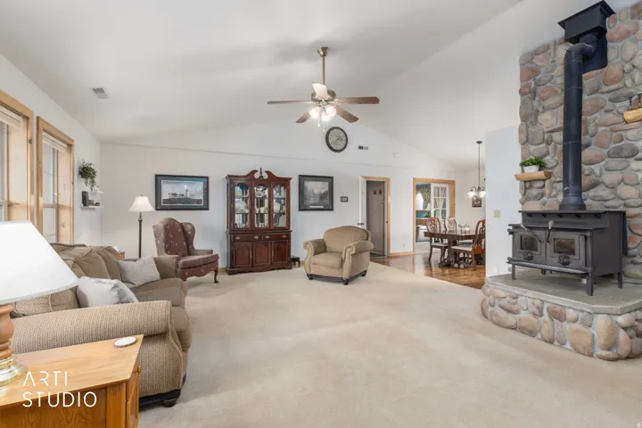Vaulted ceiling with ceiling fan, large stone-clad wood-burning stove, wood-finish window trim, light-colored carpeting, and wood-finish flooring in the dining area