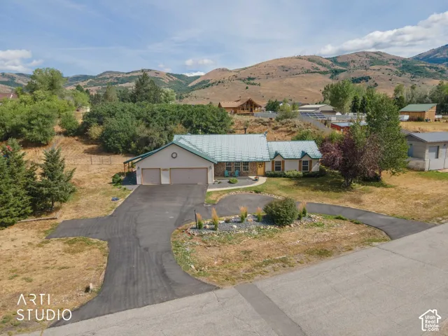 Two-car garage with overhead doors, asphalt driveway, mature landscaping, mountain backdrop, and light-toned siding