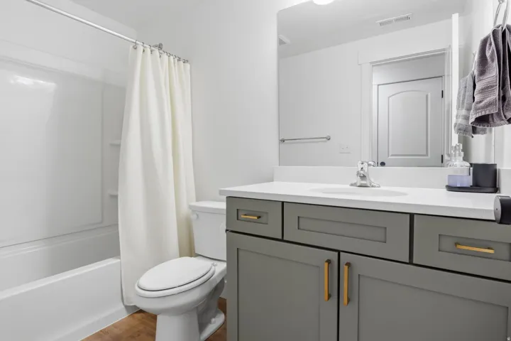 White bathtub with an integrated shower, grey vanity with gold-tone hardware, white countertop, and wood-finish flooring