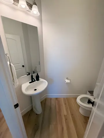 Pedestal sink with a matte black faucet, wall-mounted mirror, wood-finish flooring, and white walls
