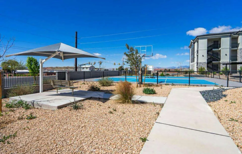 Outdoor amenity area featuring a basketball court, shaded seating, concrete pathways, decorative gravel, and a multi-story building exterior with balconies