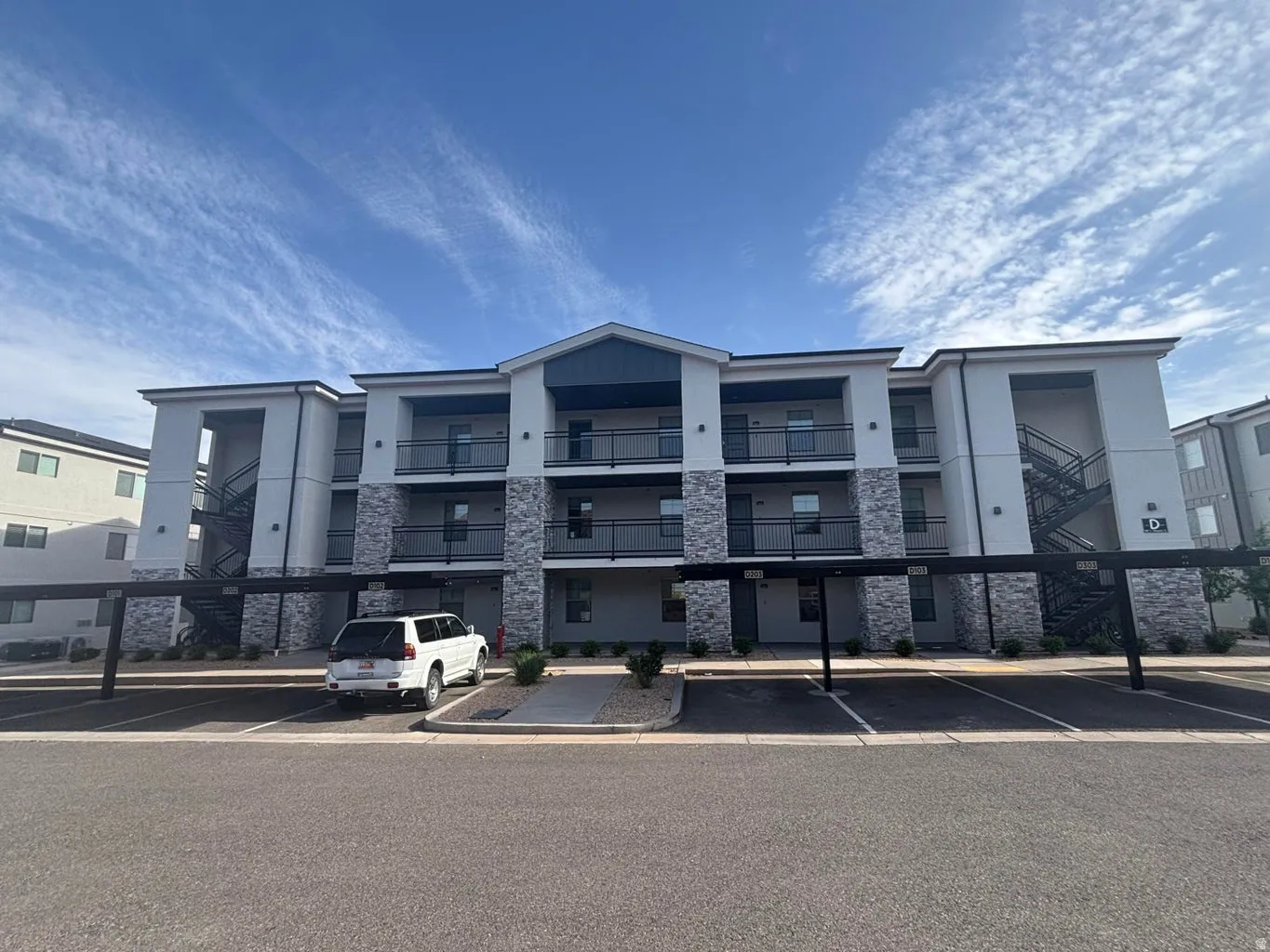 Contemporary multi-story building featuring stone veneer accents, individual balconies with black railings, and covered parking spaces