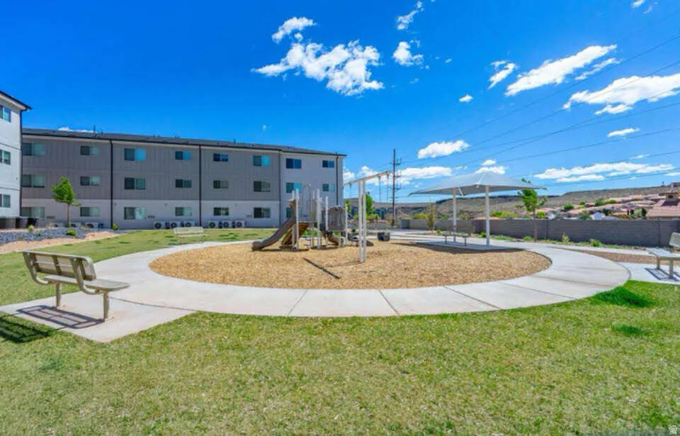 Central green space with manicured lawn, playground area with wood chip surfacing, concrete pathways, and a covered shade structure
