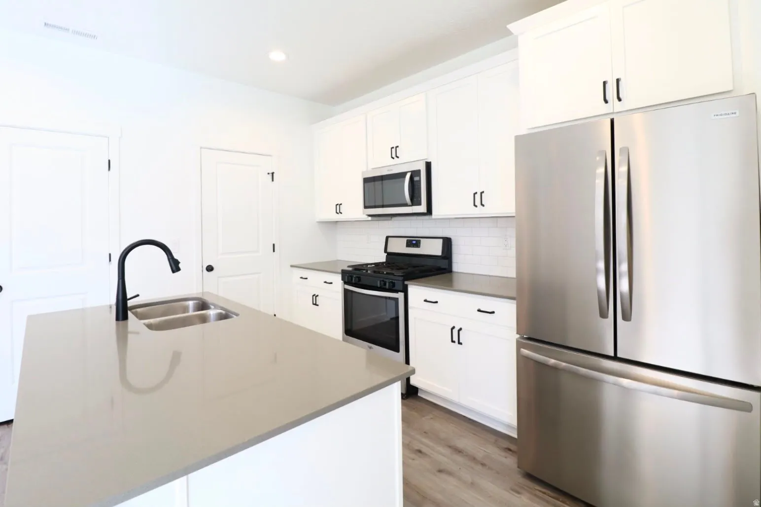 Kitchen featuring stainless steel appliances, white cabinetry, a kitchen island with a double basin sink, light-colored countertops, and wood-finish flooring